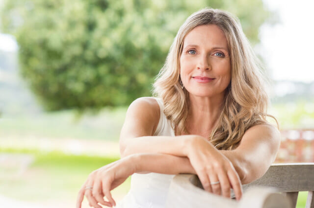 Happy senior woman relaxing on bench in the lawn. Close up face of a mature blonde woman smiling and looking at camera. Retired woman in casuals sitting outdoor in a summer day.