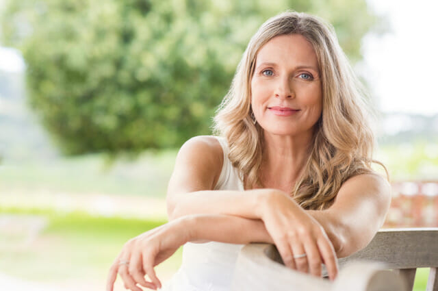 Happy,Senior,Woman,Relaxing,On,Bench,In,The,Lawn.,Close Happy senior woman relaxing on bench in the lawn. Close up face of a mature blonde woman smiling and looking at camera. Retired woman in casuals sitting outdoor in a summer day.